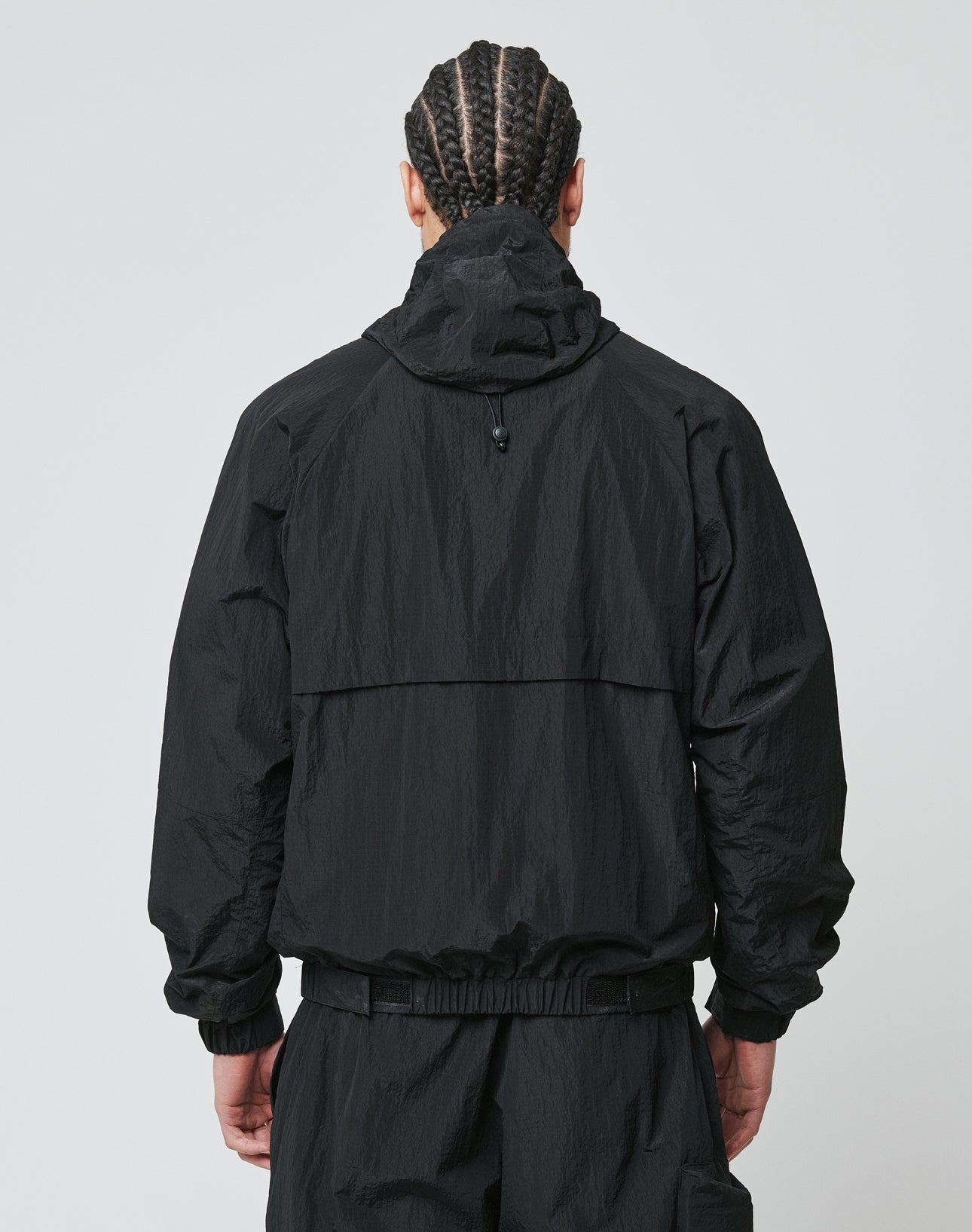 A person with braided hair stands with their back to the camera, wearing a black LIVE FAST DIE YOUNG Live Fast Nylon Jacket with seitliche Eingrifftaschen and oversized black pants against a plain light background.