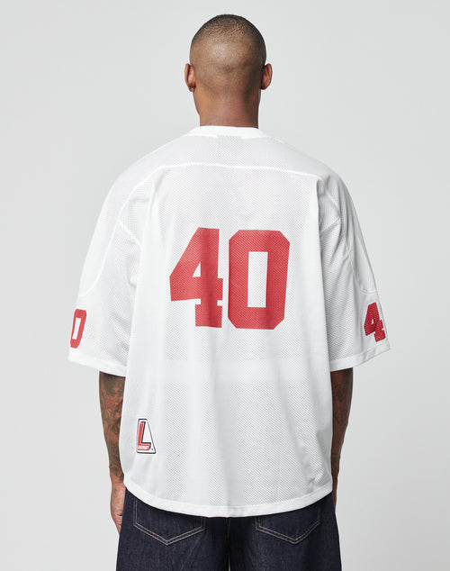 A person with a shaved head is seen from behind wearing an oversize white LFDY Mesh Jersey by LIVE FAST DIE YOUNG, featuring red number 40 on the back and sleeves, paired with dark jeans. The background is plain light gray.