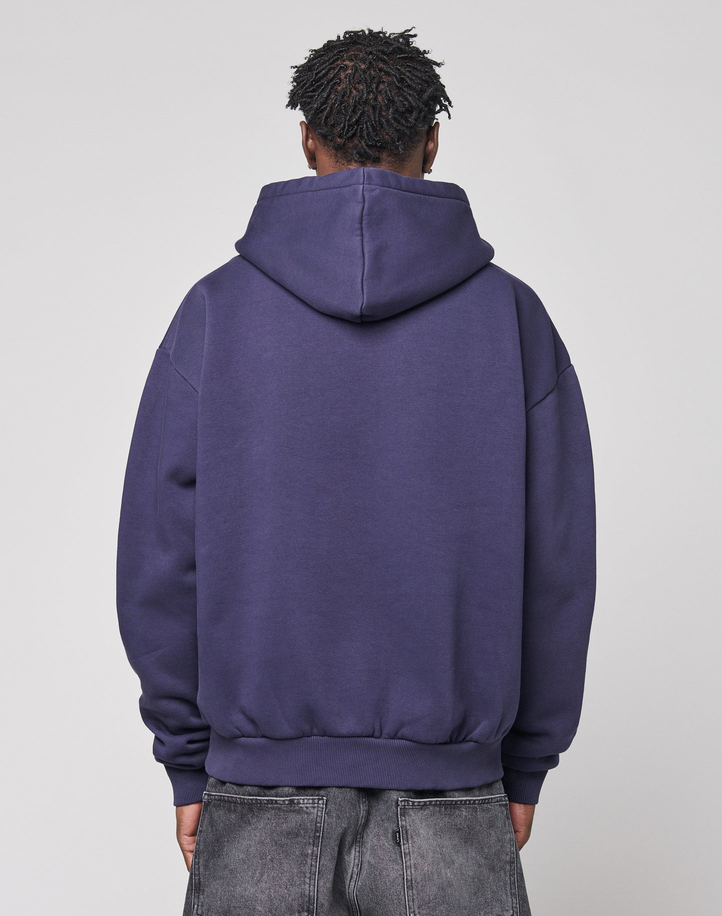 Someone is seen from behind, wearing the LIVE FAST DIE YOUNG Live Fast Stallion Hooded in purple (Oversize Fit) and faded gray jeans, set against a plain light gray background.