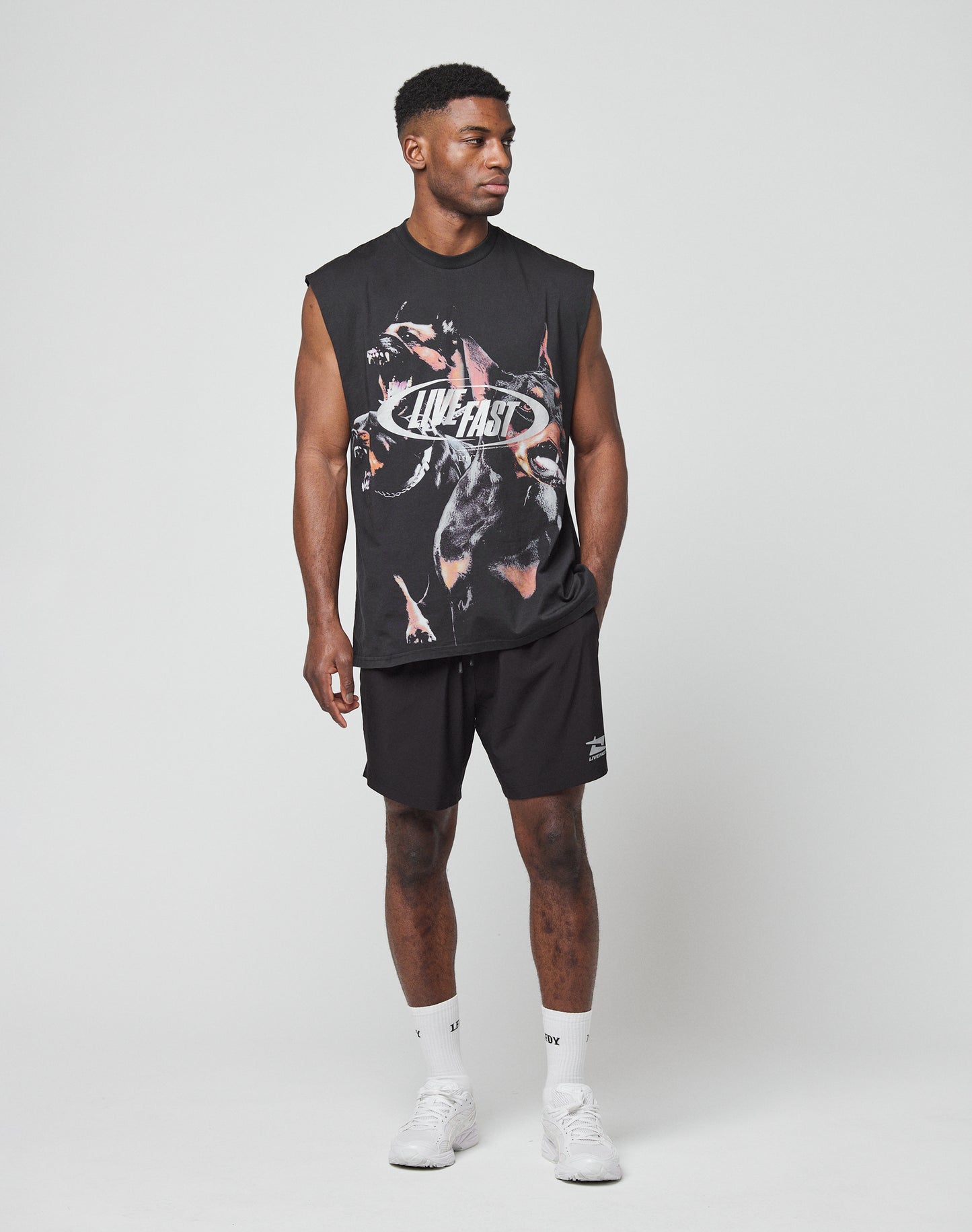 A man stands against a plain background wearing the LFDY Canine Muscle Shirt by LIVE FAST DIE YOUNG, paired with black shorts, white socks, and white sneakers. He looks to the side with one hand in his pocket.