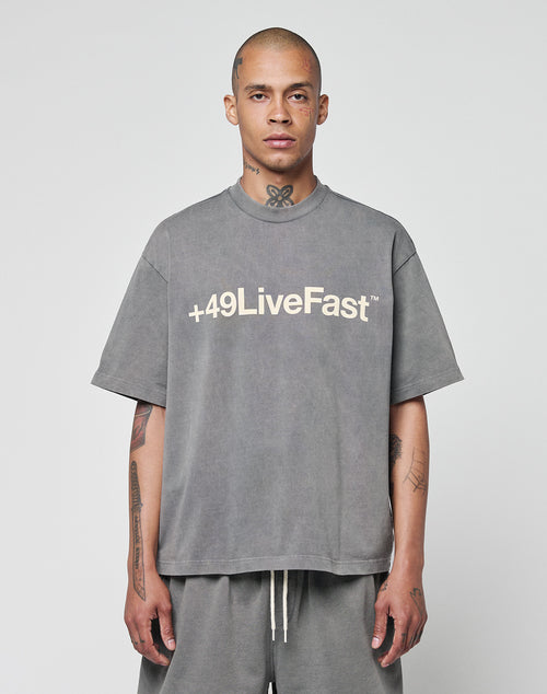 A tattooed, shaved-head individual wears the oversize-fit Basic +49 Tee by LIVE FAST DIE YOUNG, in gray 100% Baumwolle with a bold +49LiveFast front print, against a plain light background.