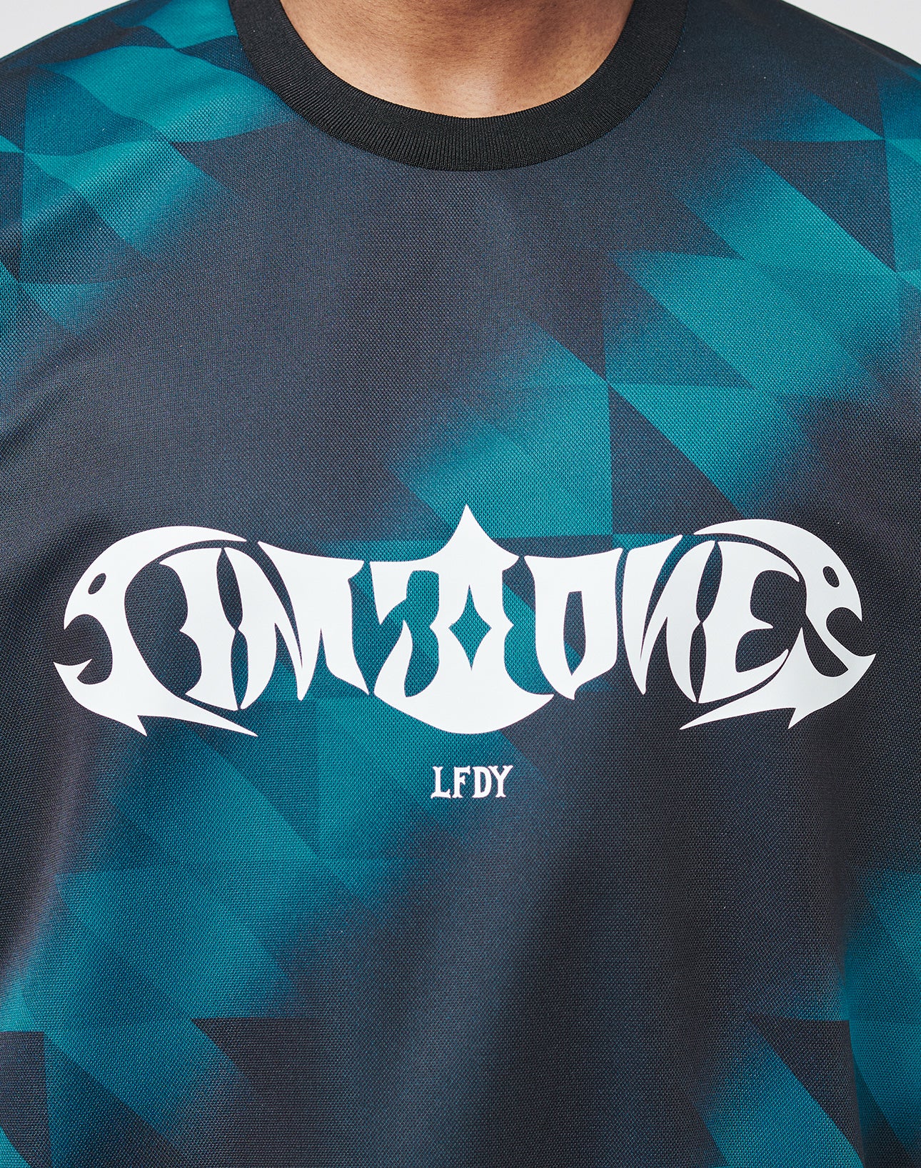 A close-up of a person wearing the LIVE FAST DIE YOUNG Live Fast Jersey Tee, a regular fit blue and black geometric-patterned polyester shirt with bold IMPZONE across the chest and LFDY printed below.