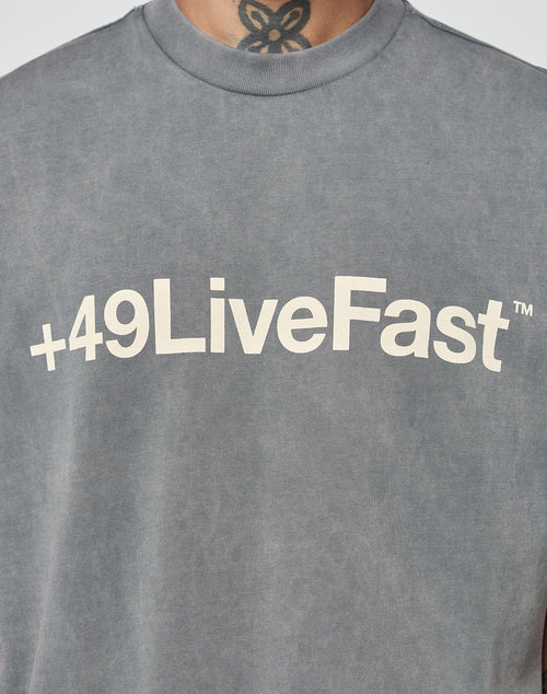A person wears the oversize Basic +49 Tee by LIVE FAST DIE YOUNG in faded gray, made of 100% Baumwolle, featuring a large +49LiveFast™ front print; a black neck tattoo is visible.