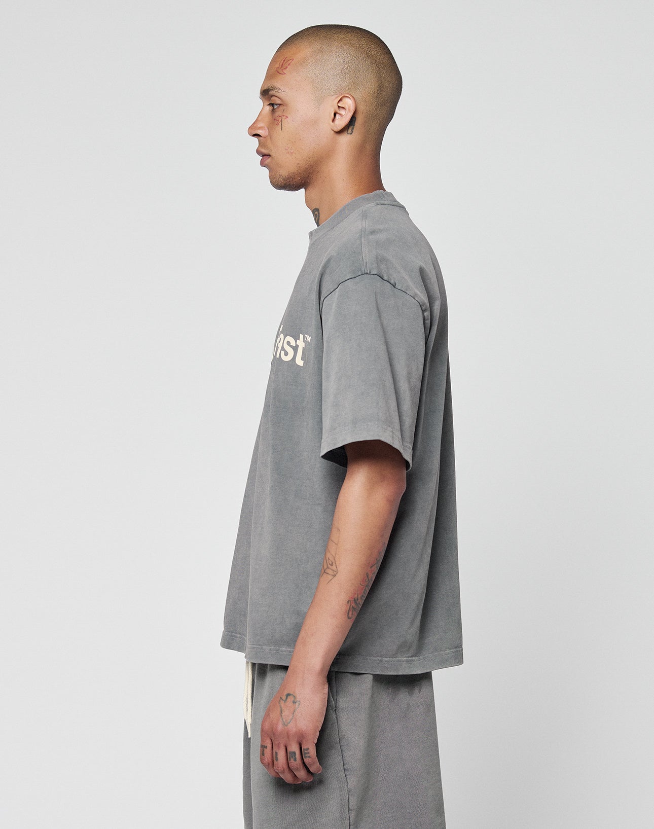 A man with a shaved head and tattoos stands in profile, wearing the LIVE FAST DIE YOUNG Basic +49 Tee—an oversize, faded gray 100% Baumwolle shirt with a partial white front print—paired with matching gray shorts against a plain background.