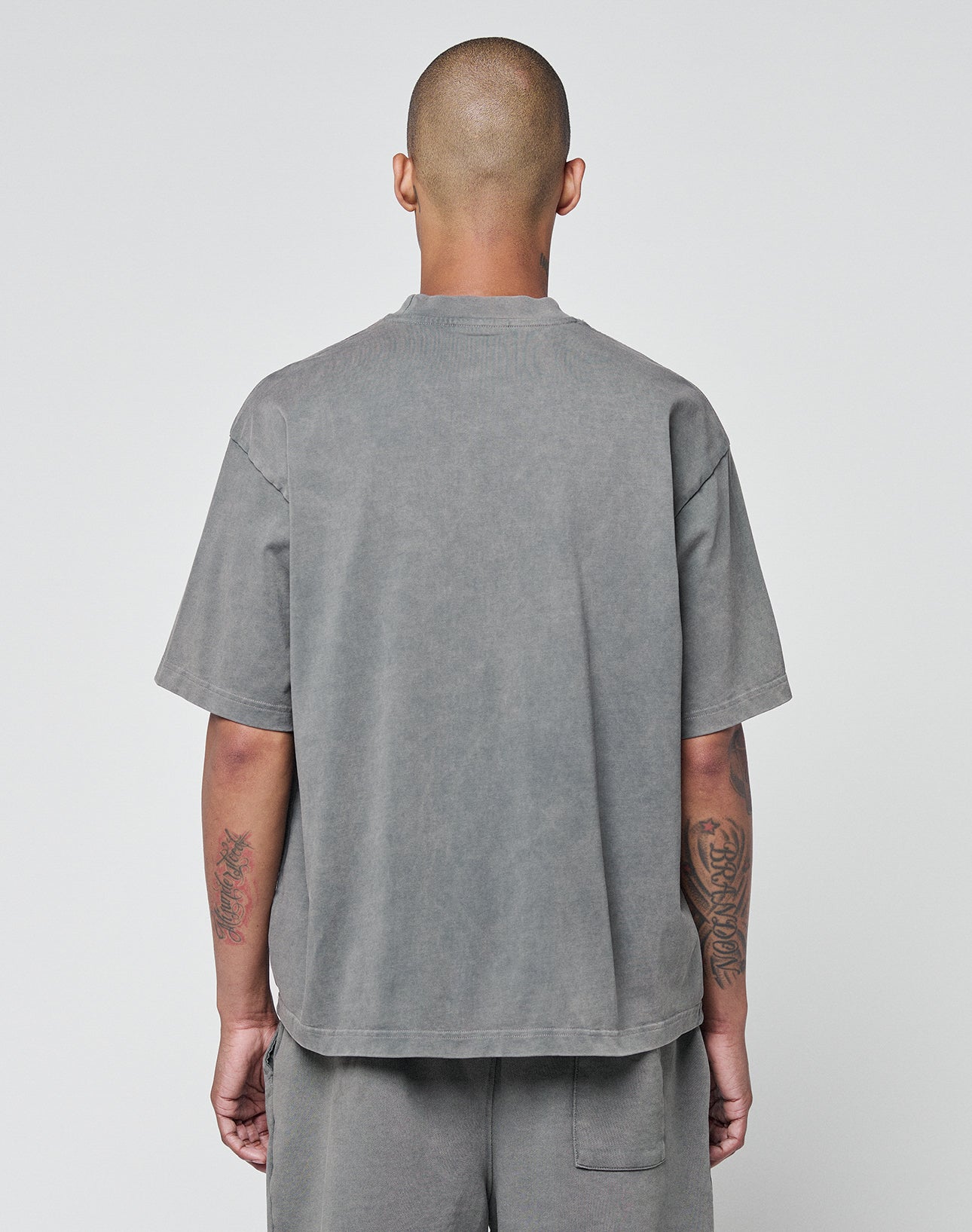 A person with a shaved head stands with their back to the camera, wearing the LIVE FAST DIE YOUNG Basic +49 Tee—an oversize fit, short-sleeved gray shirt made from 100% Baumwolle—paired with matching pants. Tattoos are visible on their arms.