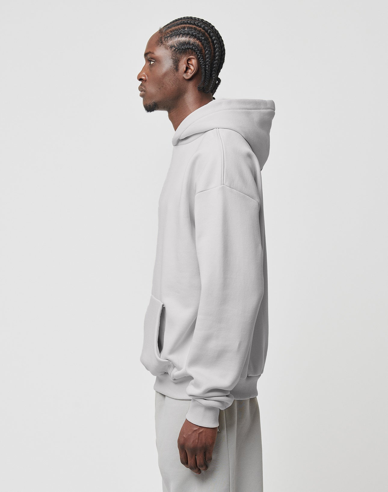 A person with braided hair stands in profile wearing a LIVE FAST DIE YOUNG Basic Blank Hooded light gray hoodie and matching pants, made from a soft cotton-polyester blend, against a plain white background.
