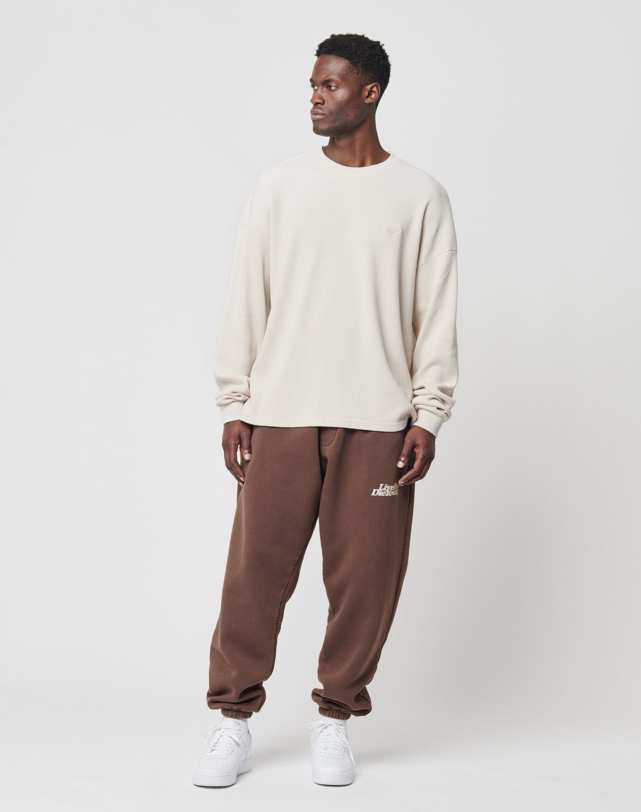A man stands against a plain white background wearing LIVE FAST DIE YOUNGs Basic Jogger in brown with a Breiter Elastikbund and white text on the left thigh, paired with a loose cream sweatshirt and white sneakers. He looks left with a relaxed posture.
