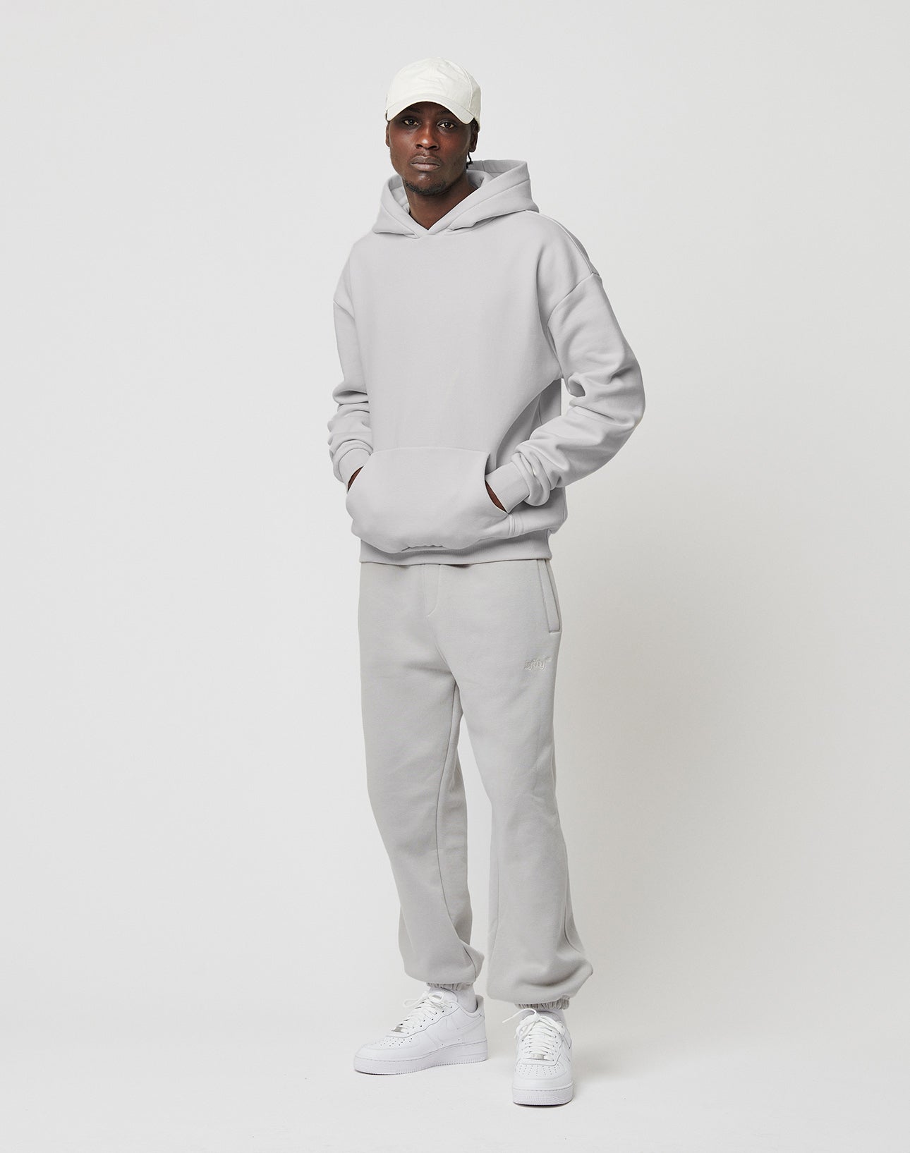A person stands against a plain white background wearing the LIVE FAST DIE YOUNG Basic Blank Hooded in light gray, matching sweatpants, white sneakers, and a white baseball cap, with hands in the hoodie pocket.