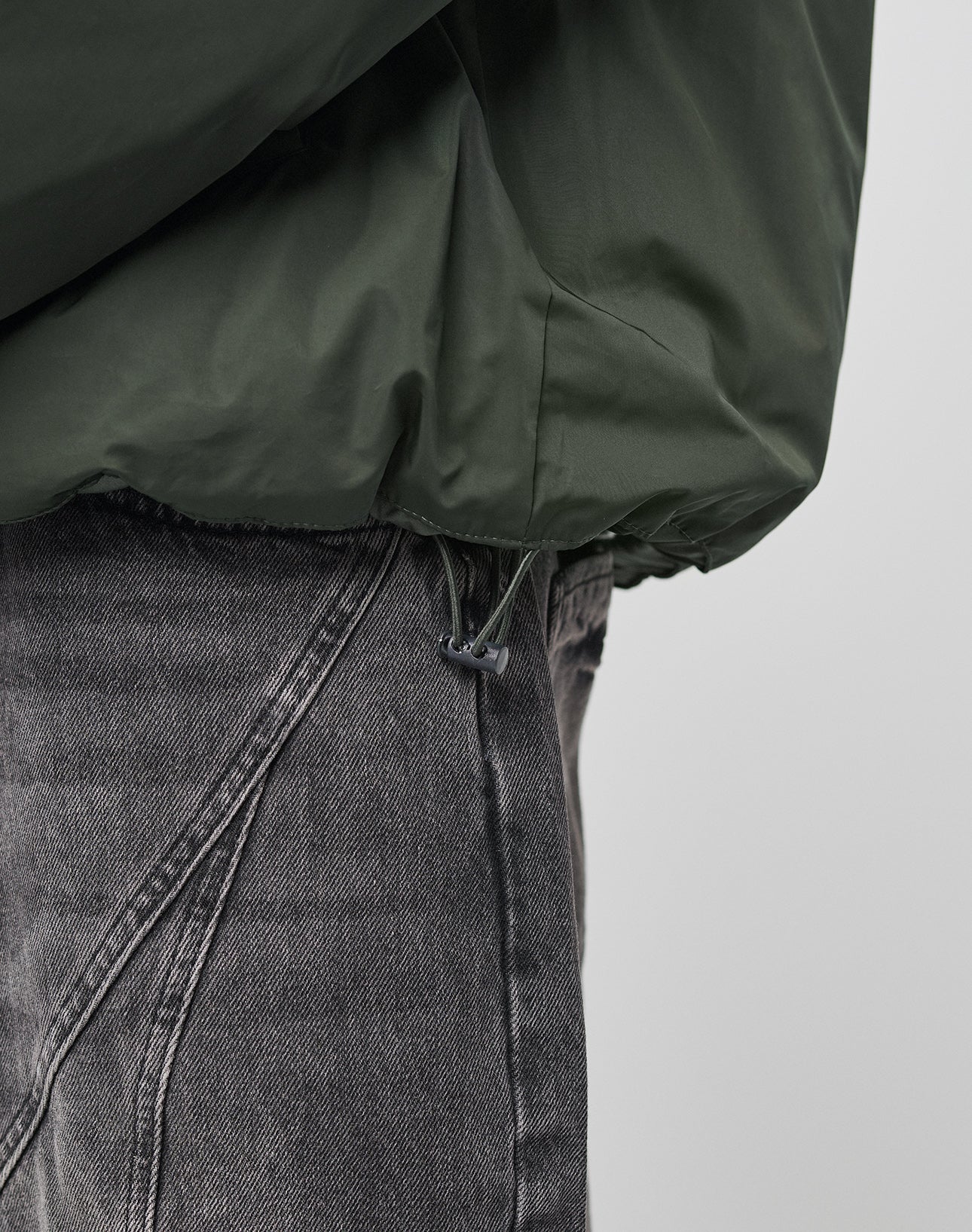 Close-up of a person wearing faded gray jeans and the LIVE FAST DIE YOUNG LFDY Puffer Jacket, featuring an adjustable drawstring hem and an oversized fit for a relaxed vibe. The background is plain and light-colored.