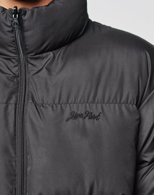 Close-up of a person wearing the LFDY Puffer Jacket by LIVE FAST DIE YOUNG. This black, oversize-fit jacket features front logo stitching, a high collar, a visible zipper, and Live Fast embroidered on the left chest.