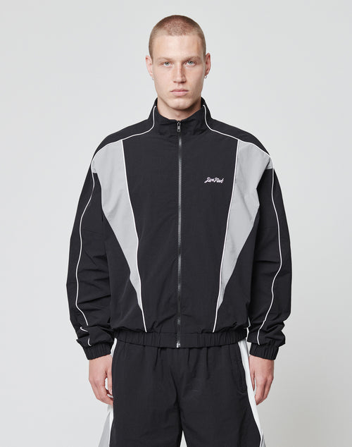 A man with short hair wears the LIVE FAST DIE YOUNG Basic Nylon Trackjacket in black and gray with seitliche Eingrifftaschen, matching pants, and a subtle Logo-Stick. He stands against a plain white background, expression neutral.
