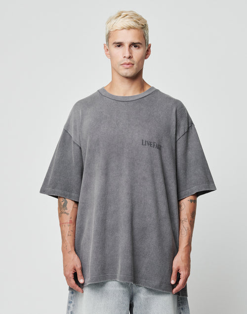 A man with short, bleached blond hair wears the LFDY Loyal Tee by LIVE FAST DIE YOUNG—an oversize fit, faded gray shirt with LIVE FAST text on the chest—and light blue jeans. He stands against a plain light background, looking serious.