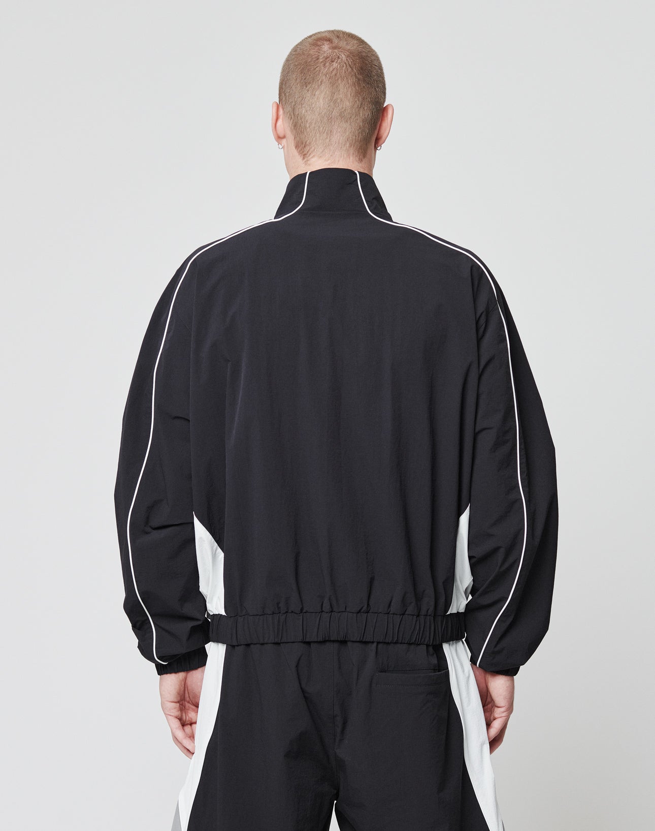 A person with short hair faces away from the camera, wearing a black and white LIVE FAST DIE YOUNG Basic Nylon Trackjacket with white piping and panels. The plain, light-colored background highlights the sporty design.