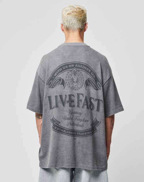 A person with short blond hair wears the LFDY Loyal Tee by LIVE FAST DIE YOUNG, a faded gray 100% Baumwolle oversize T-shirt with a large LIVE FAST graphic on the back, paired with light-wash denim shorts against a plain backdrop.