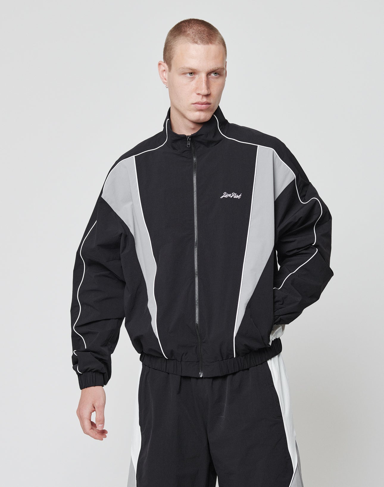 A young man with short hair wears the LIVE FAST DIE YOUNG Basic Nylon Trackjacket in black and gray with white piping and matching pants, featuring subtle logo details, standing against a plain white background.