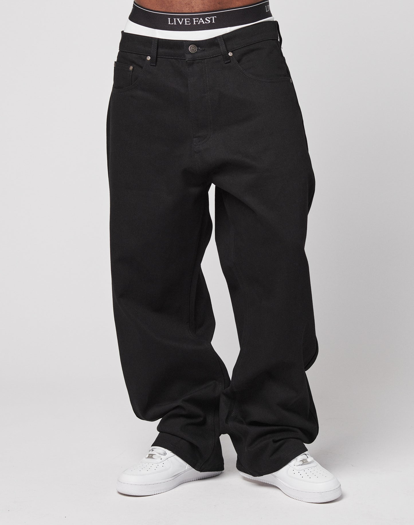 Model wears LFDY Baggy Denim by LIVE FAST DIE YOUNG in black 100% cotton, paired with white sneakers and visible black waistband featuring LIVE FAST text, set against a plain white background.