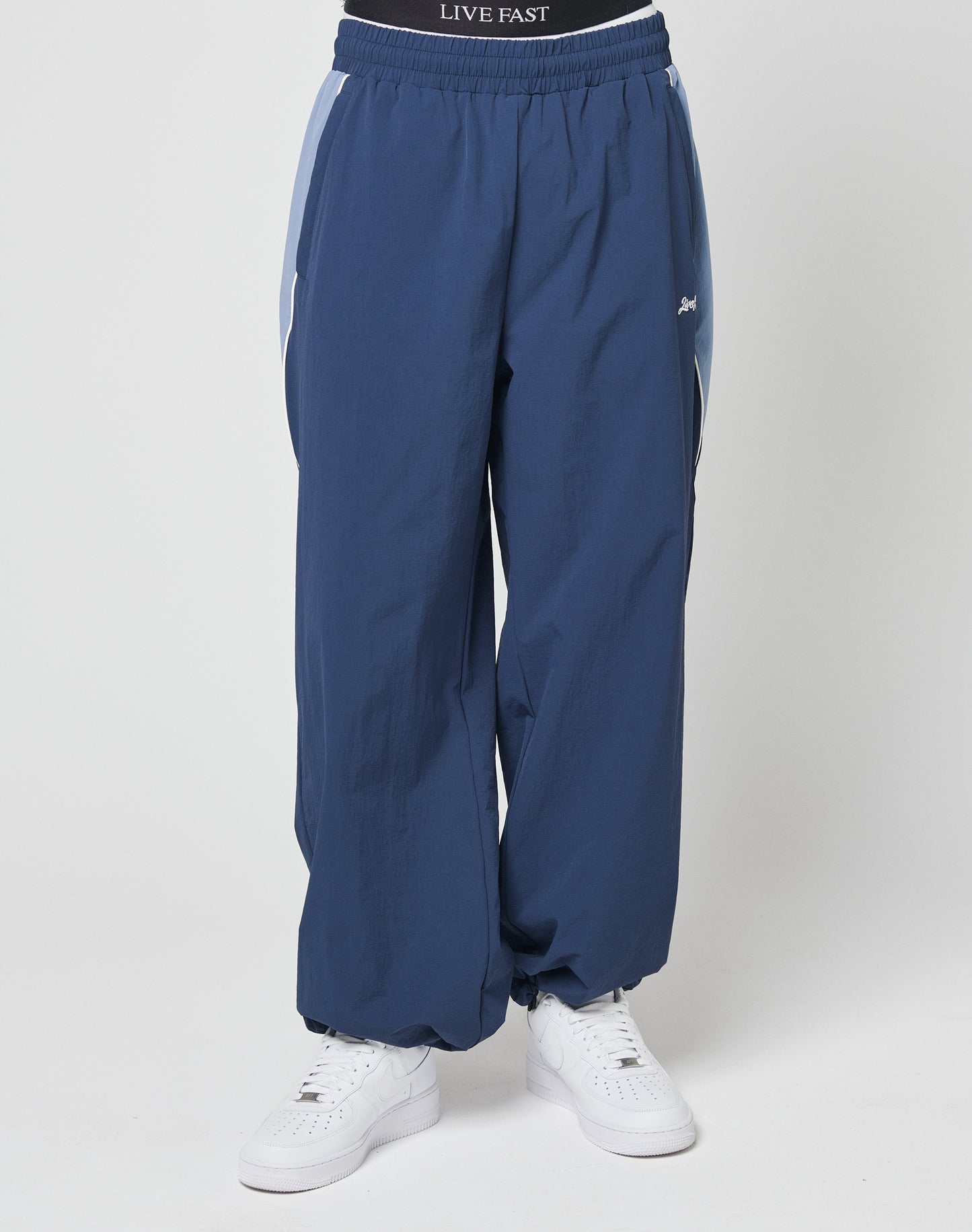 A person wearing LIVE FAST DIE YOUNG Basic Nylon Trackpants in wide fit blue with white side stripes, paired with white sneakers. The black waistband features LIVE FAST in white. Photo is cropped from the waist down.