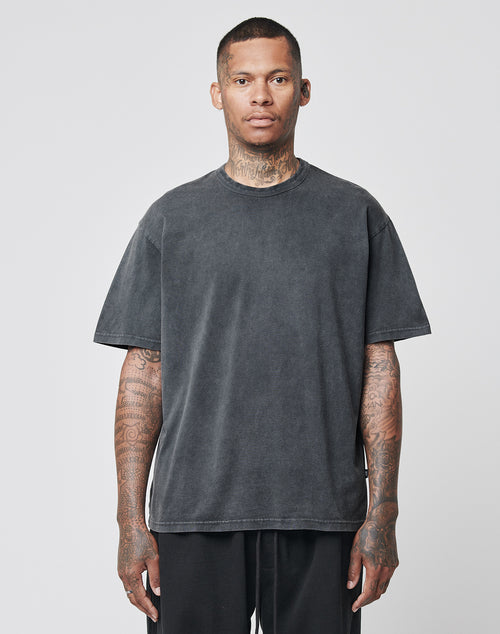 A tattooed person wears the LIVE FAST DIE YOUNG LFDY Blank Tee, a loose dark grey T-shirt aus 100% Baumwolle in Vintage-Waschung, and black regular fit pants, standing before a plain light grey background and looking straight ahead.