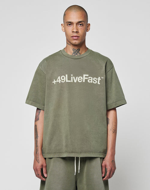A person with a shaved head and tattoos wears a LIVE FAST DIE YOUNG Basic +49 Tee in olive green, made from 100% Baumwolle, featuring +49 LiveFast on the front, standing against a plain light background.
