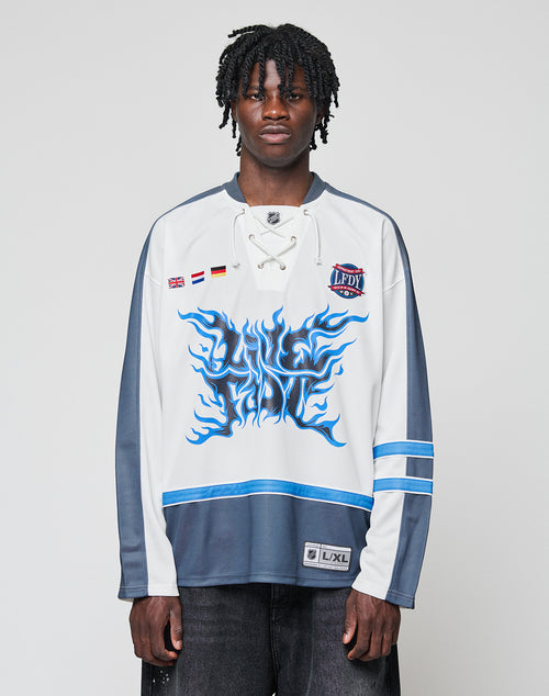 A man with short dreadlocks wears the LIVE FAST DIE YOUNG LFDY Flames Jersey—an oversized white and gray hockey top with blue flame graphics, front print details, and stripe patches—standing against a plain light background.