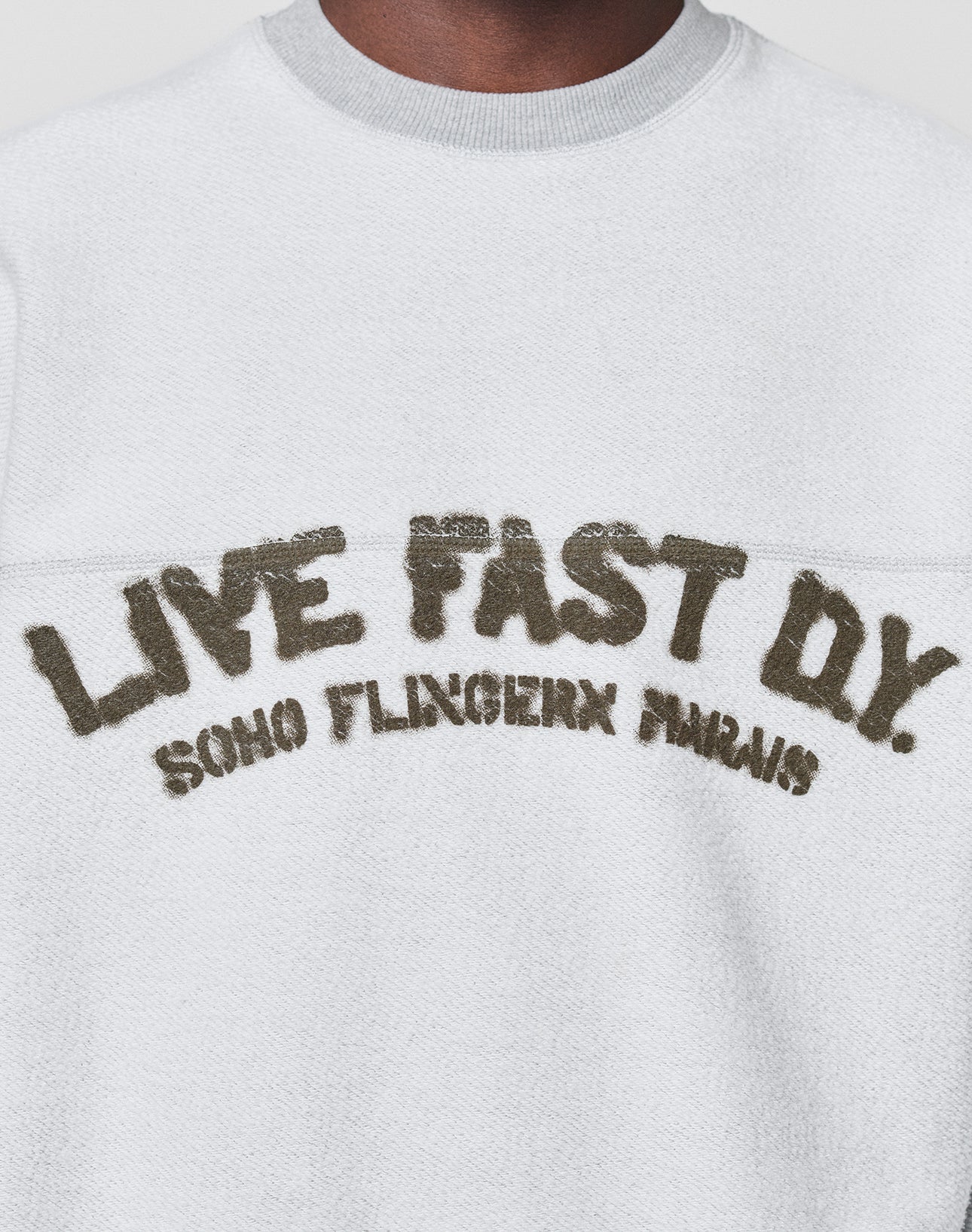 A person wears the LFDY District Sweater by LIVE FAST DIE YOUNGāan oversized white textured sweatshirt in soft cotton-polyester, featuring "LIVE FAST D*Y. SOHO FLINGERN PARIS" in large, distressed letters on the chest.