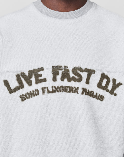 A person wears the LFDY District Sweater by LIVE FAST DIE YOUNG—an oversized white textured sweatshirt in soft cotton-polyester, featuring "LIVE FAST D*Y. SOHO FLINGERN PARIS" in large, distressed letters on the chest.