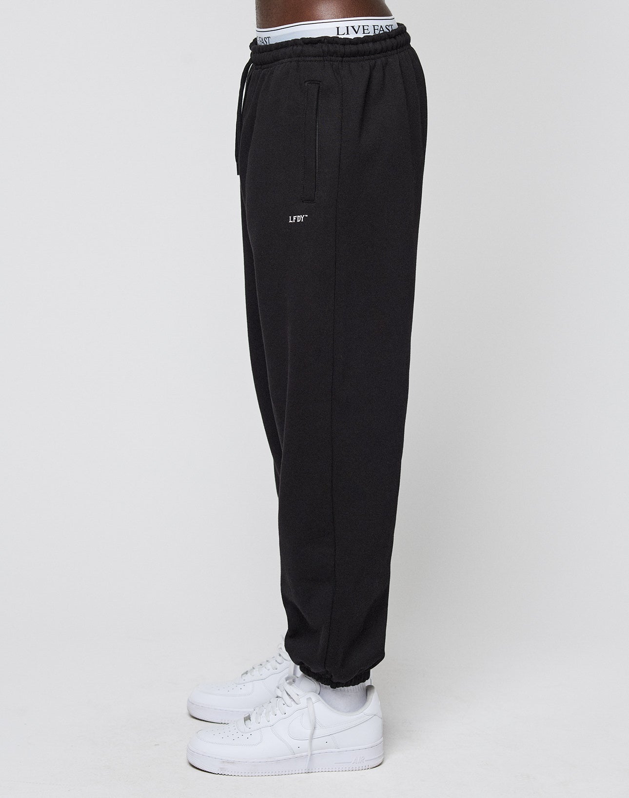 A person stands sideways in white sneakers against a plain background, wearing the LIVE FAST DIE YOUNG Basic Loose Jogger with logo print, an Elastik-Bund waistband reading LIVE FAST, and LIFT on the thigh.