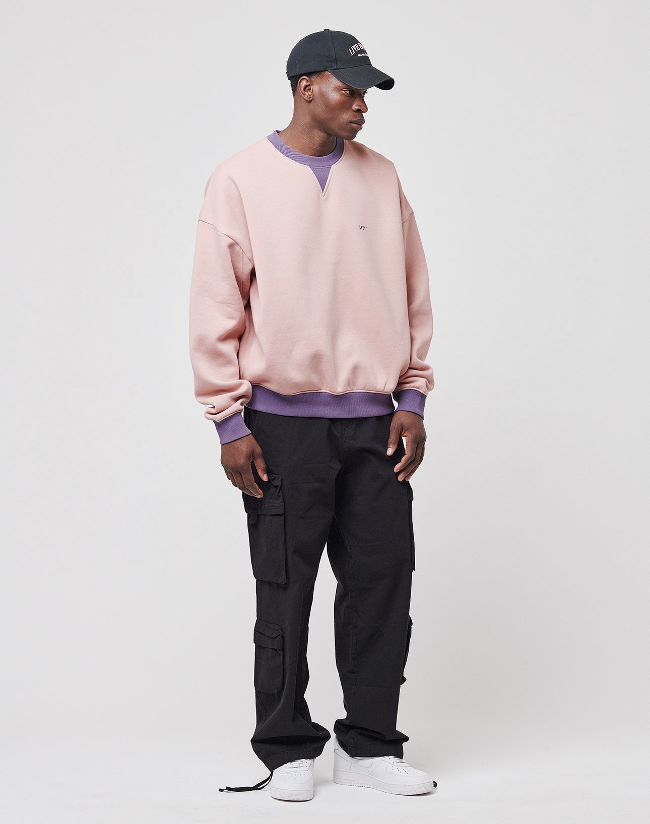 A person wearing a black cap, pale pink sweatshirt with purple accents, LIVE FAST DIE YOUNG Wide Summer Cargo pants in black ripstop, and white sneakers stands against a plain white background.