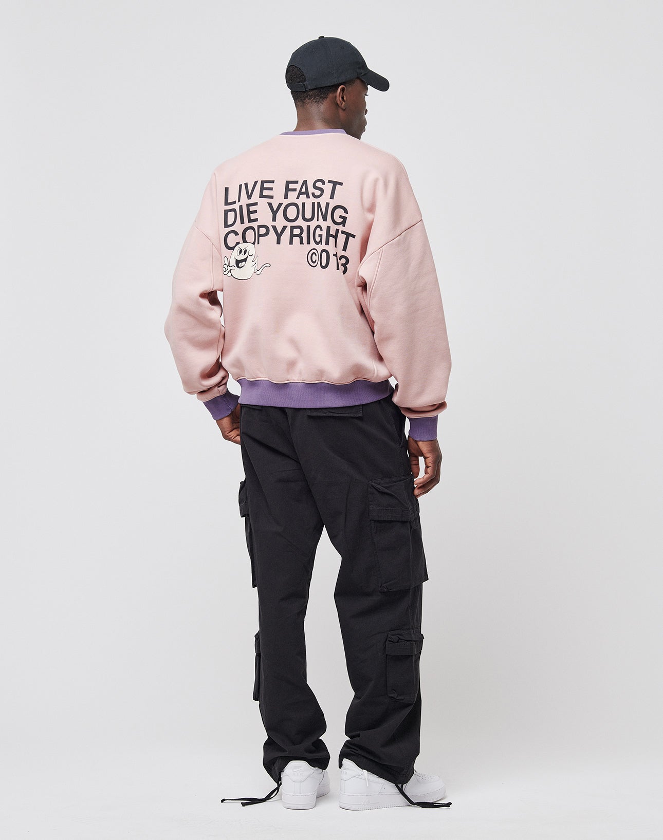 A person faces away wearing the LIVE FAST DIE YOUNG Copyright Sweater—an oversized pink sweatshirt with bold black text, paired with black cargo pants, white sneakers, a black cap, and a purple shirt visible at the sleeves.