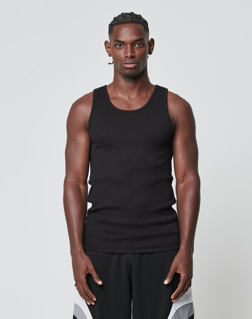 A man stands facing forward against a plain background, wearing the LIVE FAST DIE YOUNG Blank Rib Tank in slim fit black and black-and-white athletic shorts. Both feature Flag Label details and are crafted from Baumwolle. His arms are relaxed at his sides.
