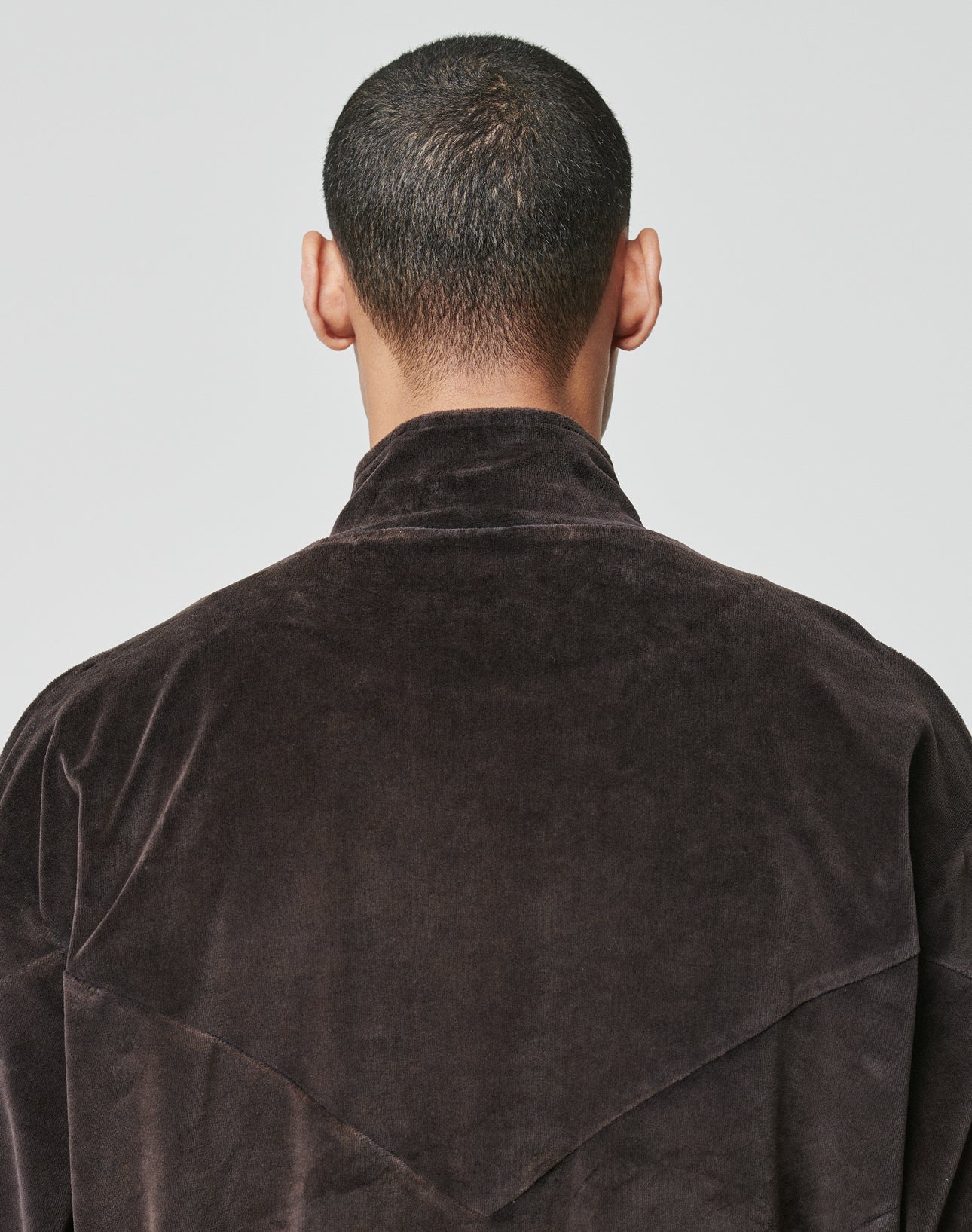 A person with short dark hair, seen from behind, wears the oversize fit LIVE FAST DIE YOUNG LFDY Velour Jacket in dark brown with a high Stehkragen and visible seam details.
