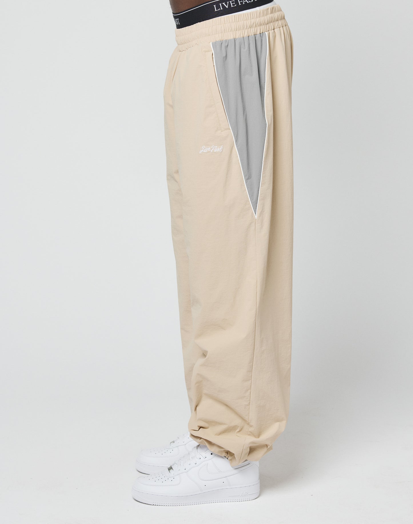 A person is wearing LIVE FAST DIE YOUNG Basic Nylon Trackpants in beige with a light gray side panel, Reißverschluss Seitentaschen, and an Elastik-Kordelzug, paired with white sneakers and standing sideways against a plain white background.