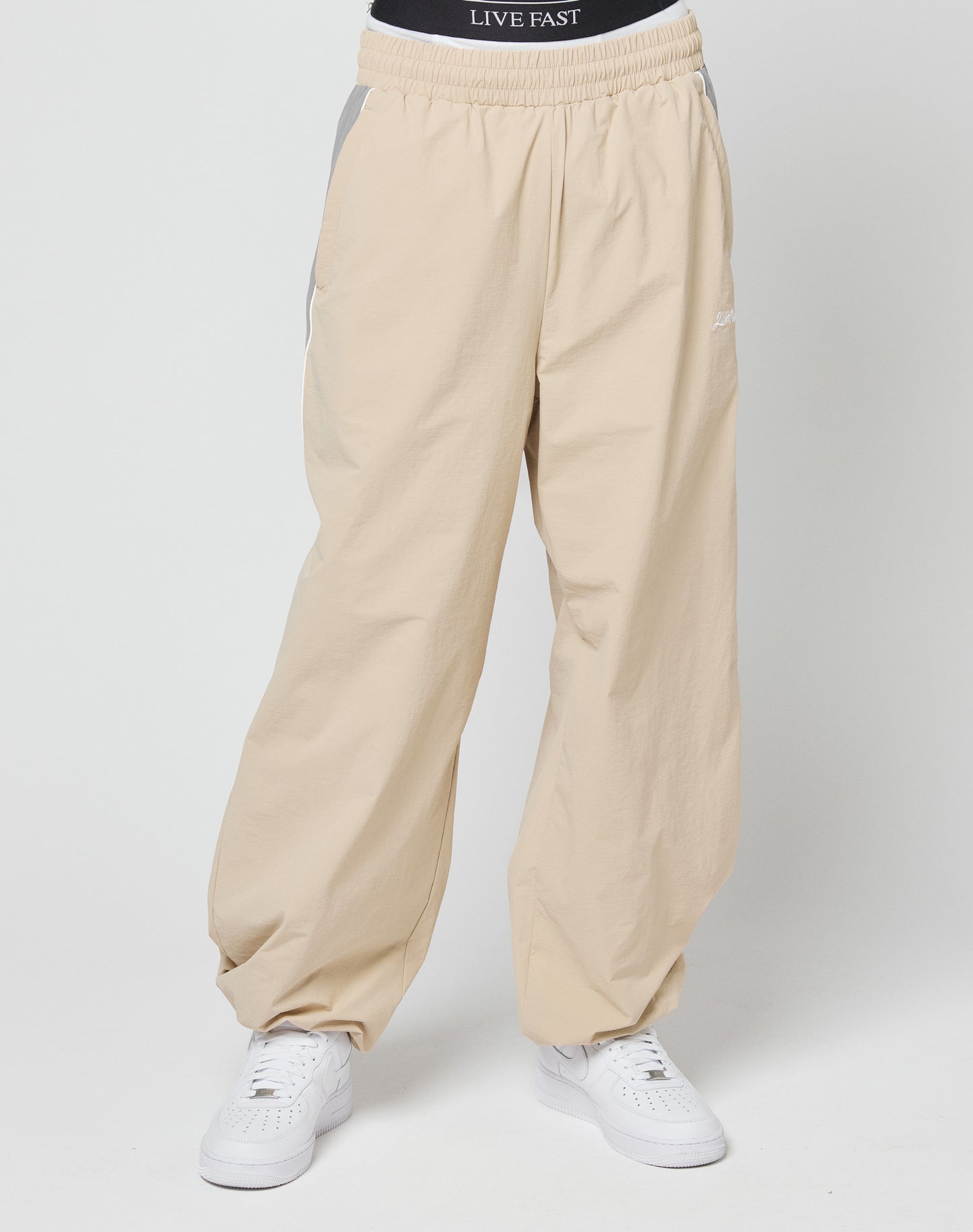 A person wears beige Basic Nylon Trackpants from LIVE FAST DIE YOUNG, featuring an elastic waistband and white sneakers. The black LIVE FAST DIE YOUNG underwear’s white logo is visible above the pants. Background is plain and light-colored.