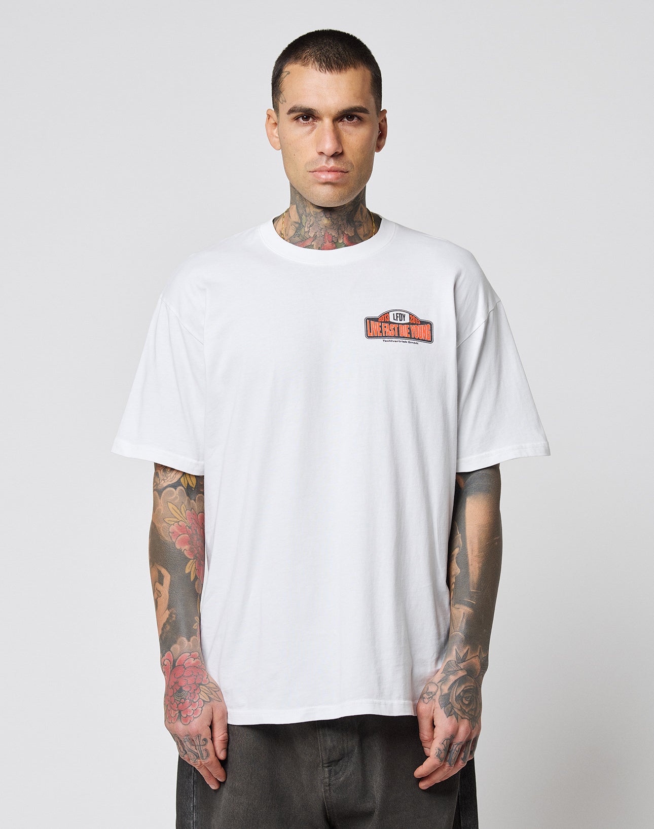 A man with tattoos on his neck and arms wears the LIVE FAST DIE YOUNG Live Fast 2013 Tee—a loose-fitting white 100% Baumwolle T-shirt with a small colored graphic and text on the left chest—standing against a plain, light background.