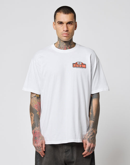 A man with tattoos on his neck and arms wears the LIVE FAST DIE YOUNG Live Fast 2013 Tee—a loose-fitting white 100% Baumwolle T-shirt with a small colored graphic and text on the left chest—standing against a plain, light background.