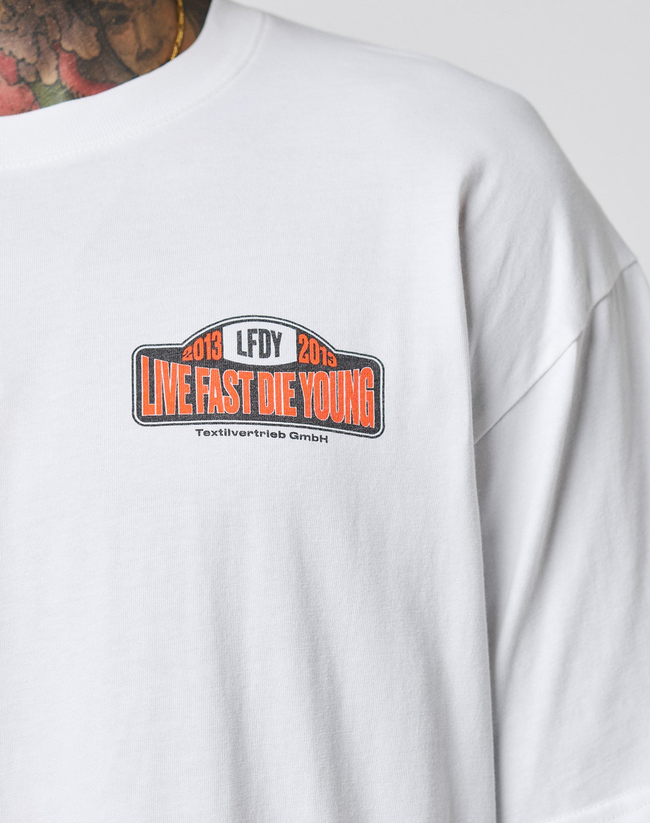 A person in the Live Fast 2013 Tee by LIVE FAST DIE YOUNG—a regular fit white shirt with LIVE FAST DIE YOUNG in bold orange, 2013 LFDY 2013, and Textilvertretieb GmbH on a chest patch. Part of a tattooed neck is visible.