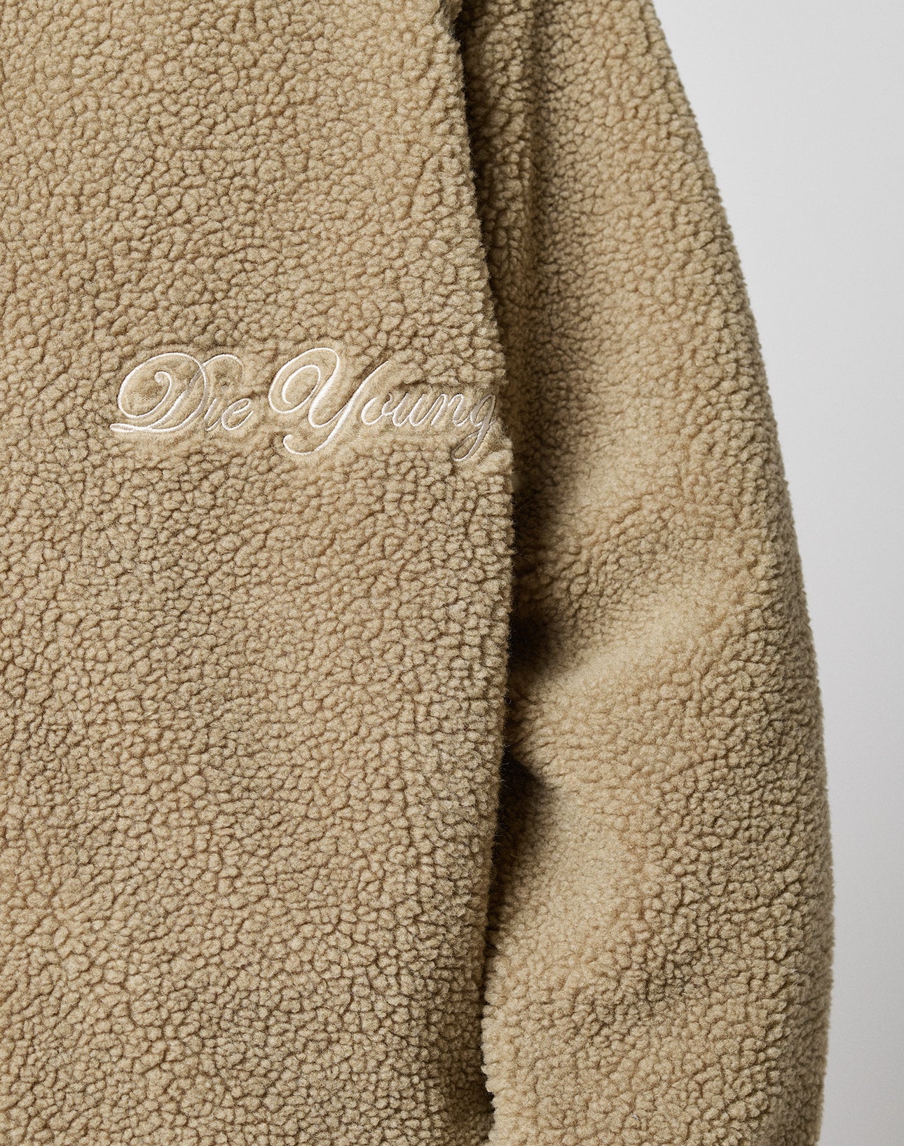 Close-up of the LFDY Teddy Jacket by LIVE FAST DIE YOUNG—a beige, fluffy, oversized jacket with a zipper and “Die Young” embroidered in white cursive on the chest.