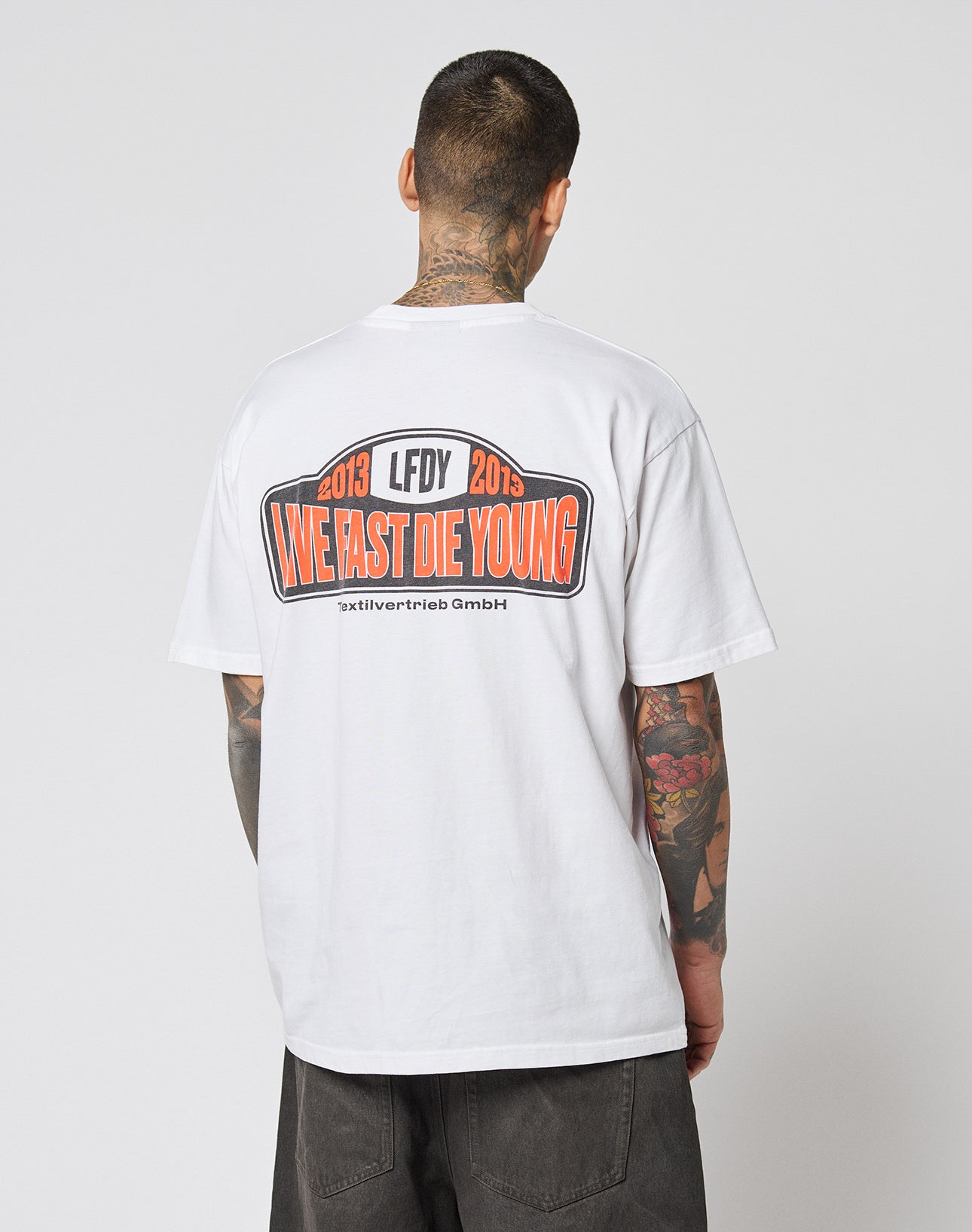 A tattooed person faces away wearing the LIVE FAST DIE YOUNG Live Fast 2013 Tee—a regular fit white 100% cotton shirt with a bold back graphic reading WE FAST DIE YOUNG, plus LFDY and 2013.