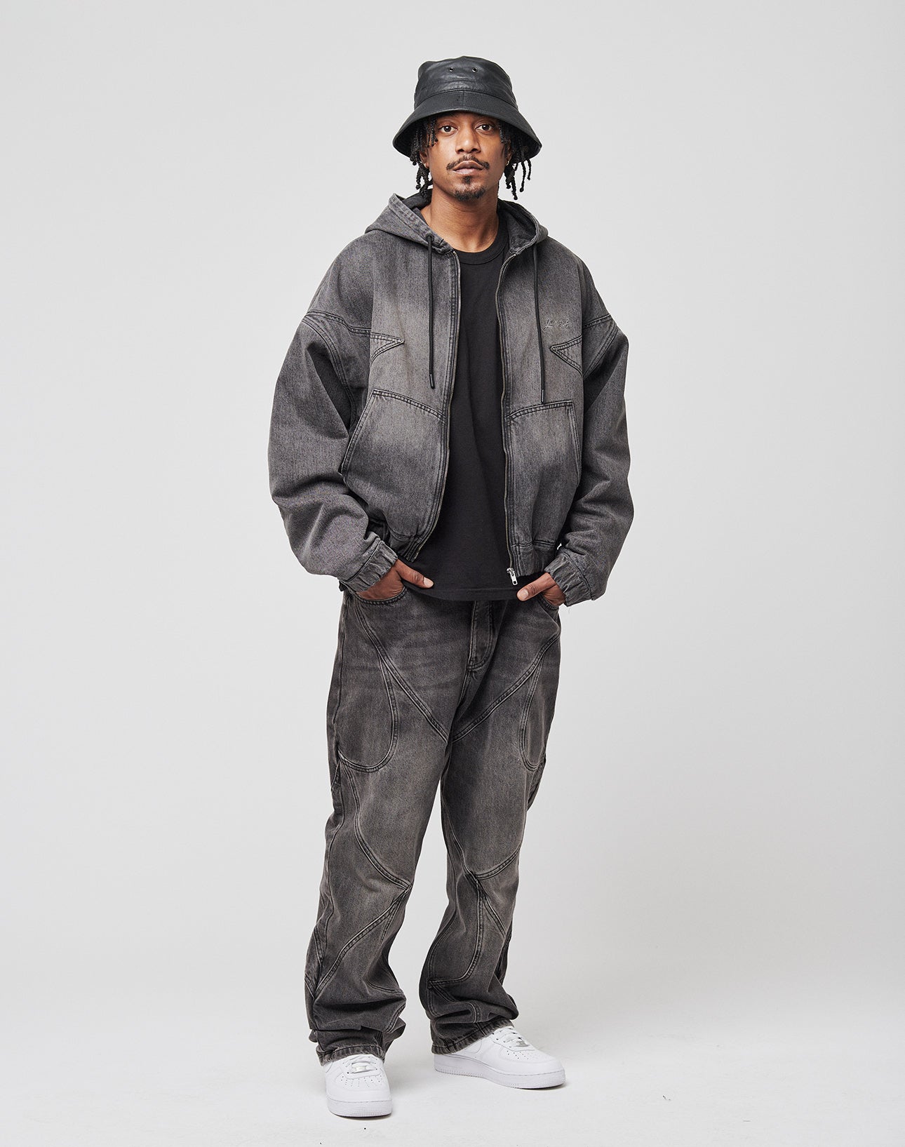 A man wears the LIVE FAST DIE YOUNG Tribal Denim Hooded Zip over a black tee, paired with gray loose jeans, a black bucket hat, and white sneakers. He stands against a plain light gray background with his hands in his pockets.