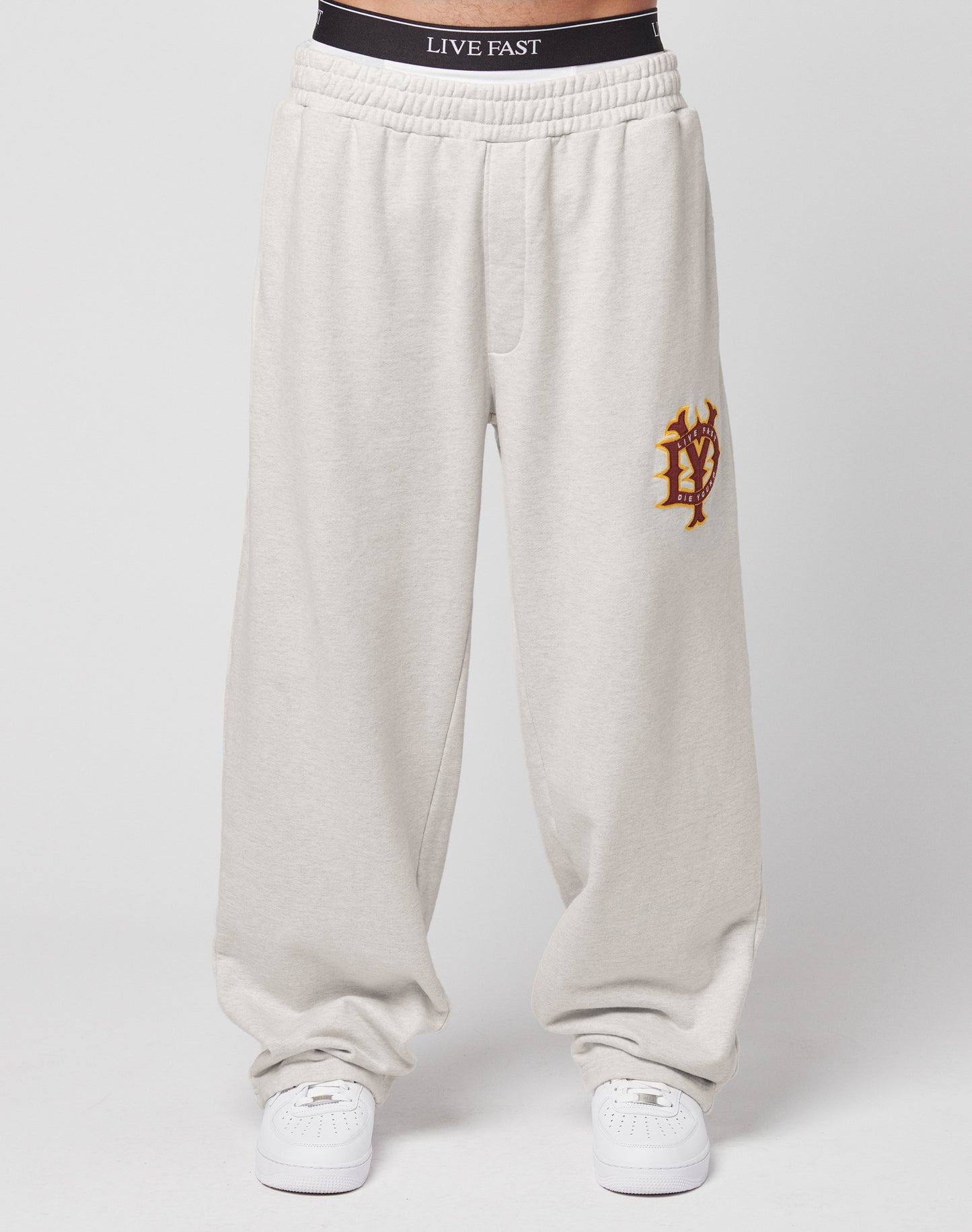 A person wears LIVE FAST DIE YOUNGs DY Jogger—an oversize fit, light gray sweatpant with wide legs, a red and yellow left-thigh emblem, and a black waistband with LIVE FAST text—paired with white sneakers against a plain white background.