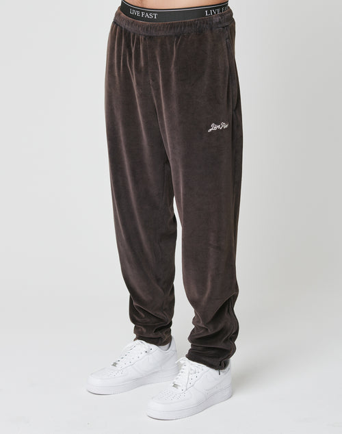 A person wears LFDY Velour Pants by LIVE FAST DIE YOUNG—loose, dark brown sweatpants with elastic waistband and LIVE FAST text, small logo on the thigh, oversize fit—paired with white sneakers against a white backdrop.