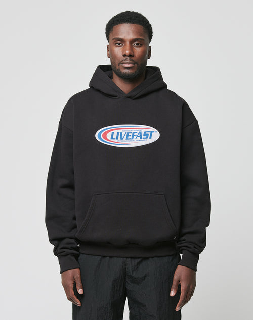 A man faces the camera wearing the Live Fast Garment Hooded by LIVE FAST DIE YOUNG, featuring a chest Live Fast logo and Flag-Label detail in premium cotton, paired with black pants against a plain light backdrop.