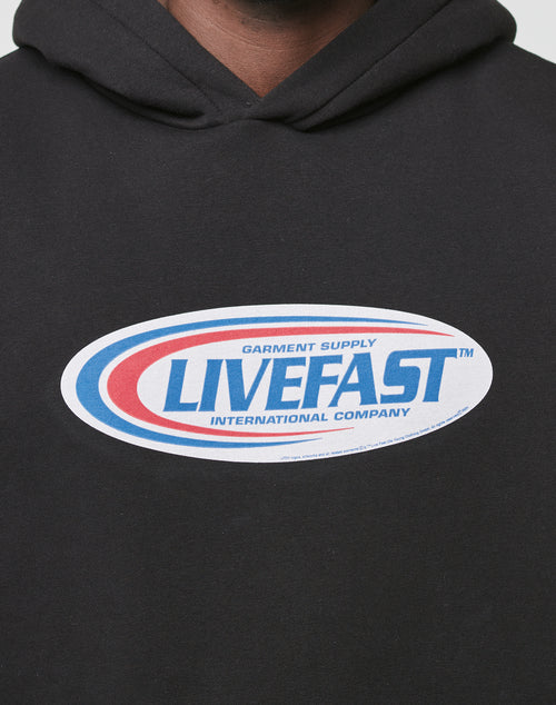 A close-up of the Live Fast Garment Hooded by LIVE FAST DIE YOUNG, an oversized black hoodie with an oval blue and red LIVEFAST Garment Supply International Company logo and a subtle flag label.