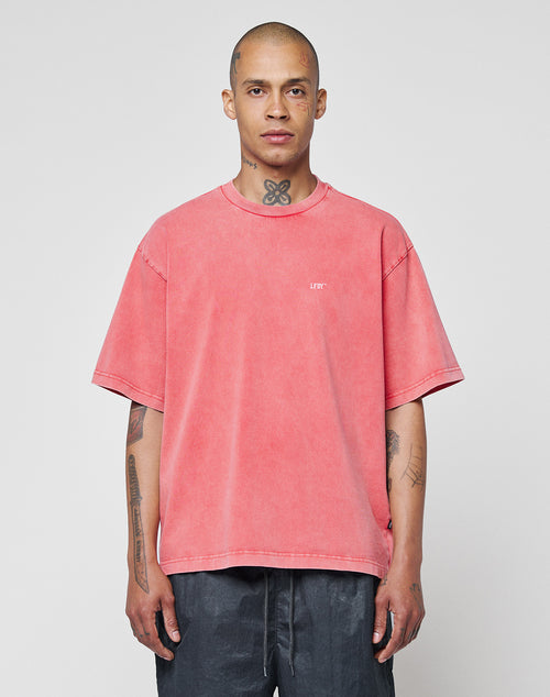 A person with a shaved head and tattoos wears the LIVE FAST DIE YOUNG Basic Evidence Tee in oversize faded pink, paired with dark pants, standing against a plain light gray background. The shirt is made from 100% Baumwolle.