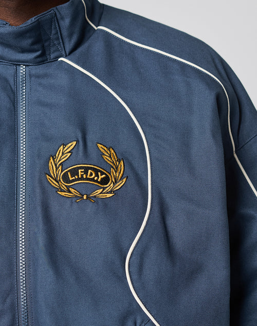 Close-up of the LFDY Canvas Jacket by LIVE FAST DIE YOUNG, featuring elastic cuffs, blue fabric, white piping, and an embroidered yellow laurel wreath with L.F.D.Y letters stitched in yellow on a black background.