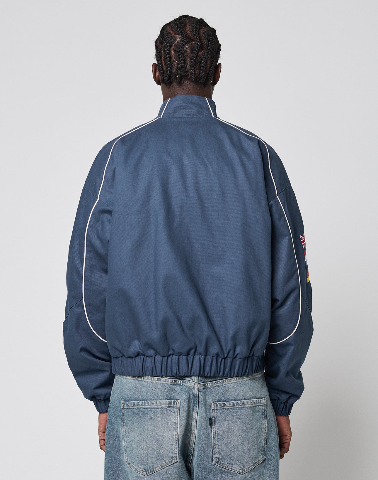 A person with braided hair is seen from behind, wearing the LFDY Canvas Jacket by LIVE FAST DIE YOUNG—blue with multilogo embroidery, white piping, elastic cuffs, and a high collar—paired with light blue jeans against a plain light background.