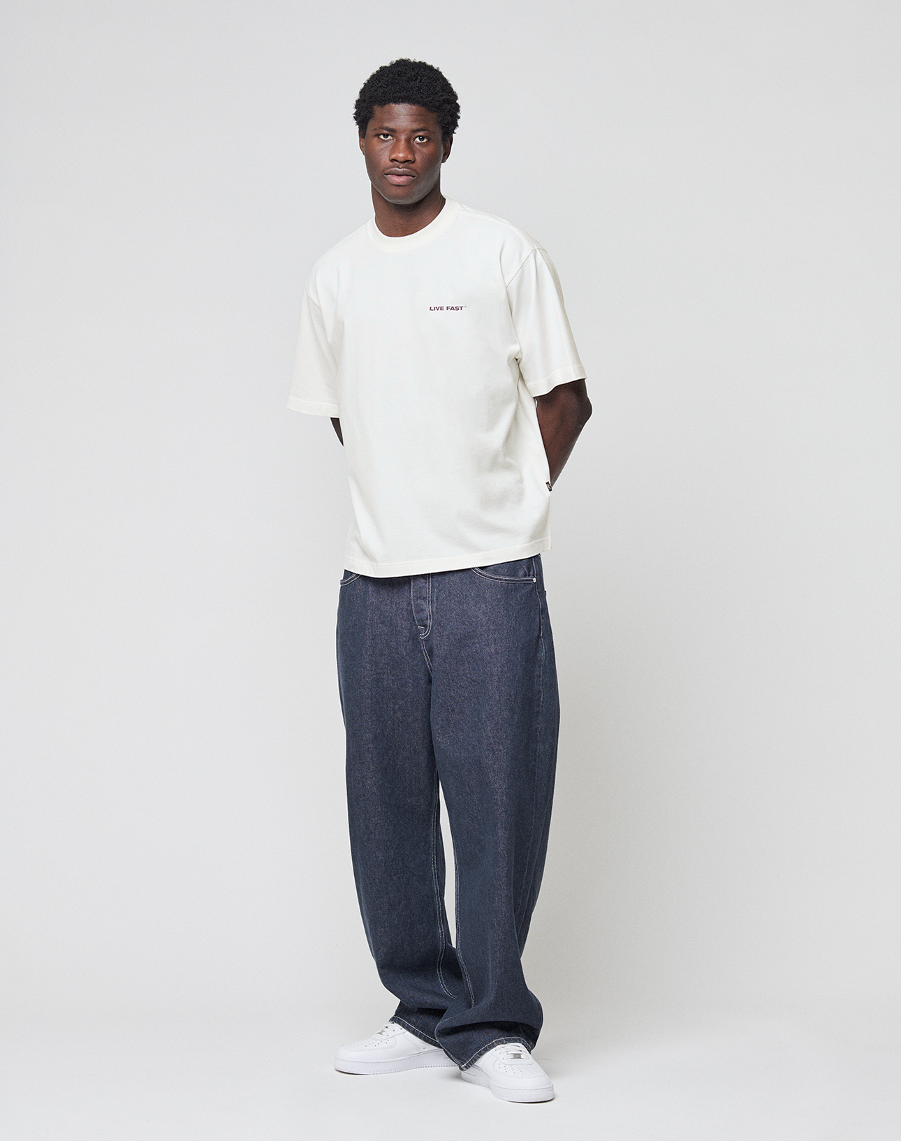 A man stands against a plain light background wearing a white LIVE FAST DIE YOUNG LFDY Grand Theft Tee, loose-fitting blue jeans, and white sneakers, with his hands behind his back.