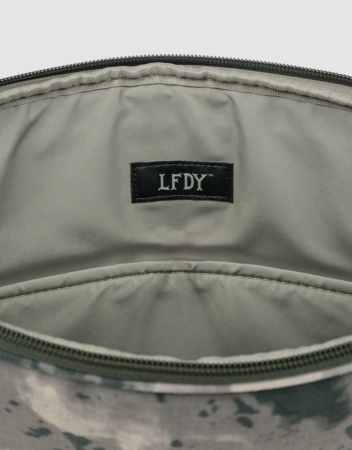 Close-up of the inside of an LFDY Laptop Case by LIVE FAST DIE YOUNG, featuring a camouflage pattern, black label with “LFDY” in white, a zip pocket, zipper closure, and light grey interior lining.