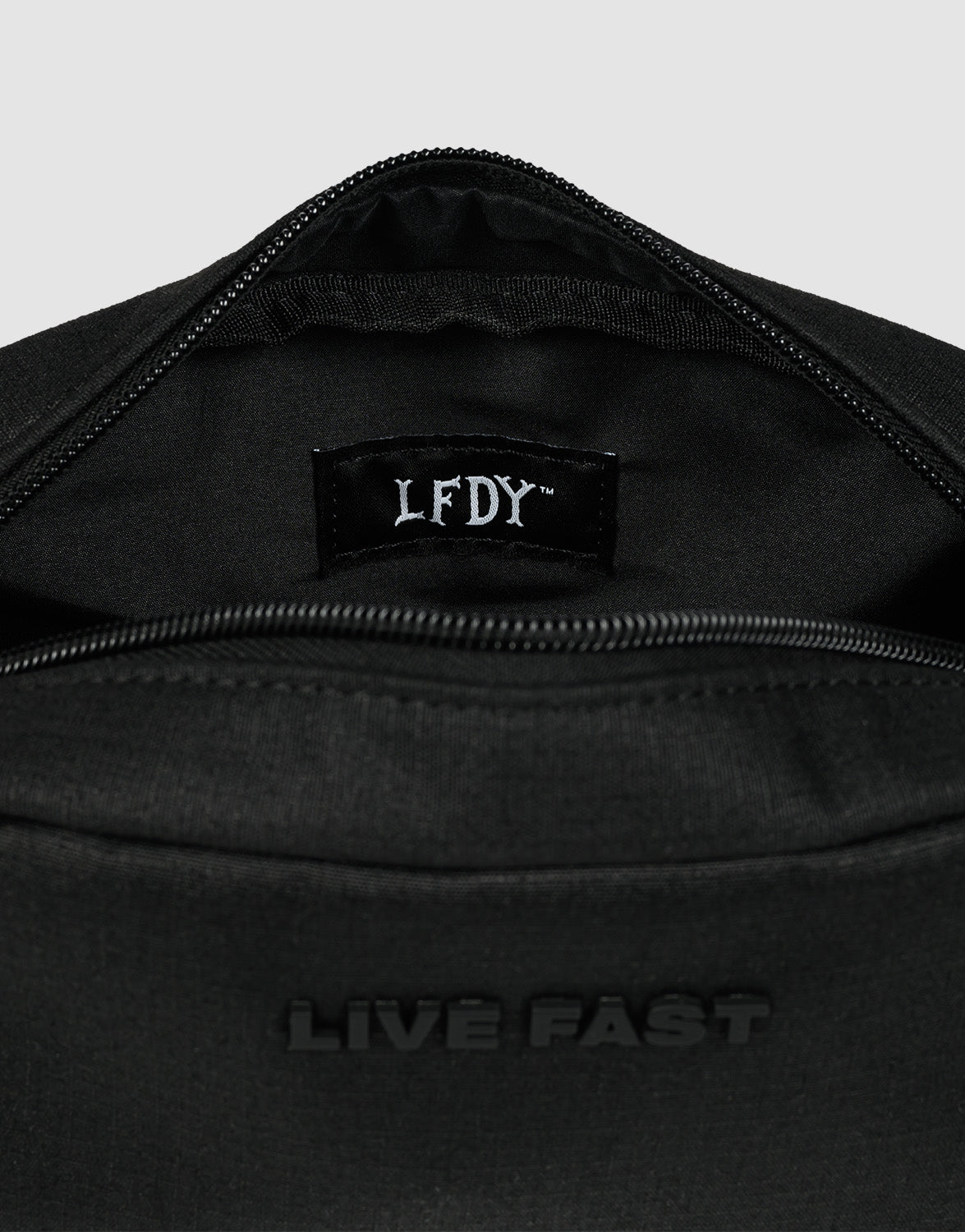 A close-up of the LFDY Amenity Kit by LIVE FAST DIE YOUNG—a black polyester zippered bag with "LIVE FAST" embossed outside and an LFDY label inside, partially open to show its inner pockets and large zip compartment.