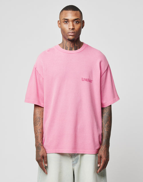 A tattooed man faces forward wearing the LFDY Loyal Tee by LIVE FAST DIE YOUNG—an oversize, loose-fit pink T-shirt made from 100% Baumwolle with LIVE FAST front print and stylish front/back designs, paired with light pants on a gray background.